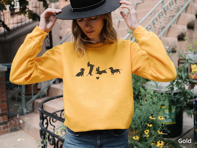 May include: A yellow sweatshirt with a graphic of four dachshund silhouettes. The dachshunds are in various poses, including sitting, standing, and running. The graphic includes a small heart.