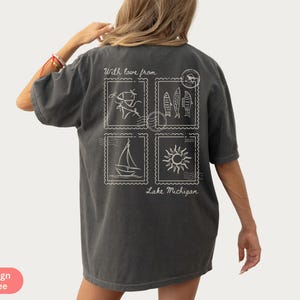 May include: Dark gray t-shirt with a white graphic design on the back. The design features four postage stamp-like images with a sailboat, fish, sun, and other elements. Text includes "With love from Lake Michigan 2023."