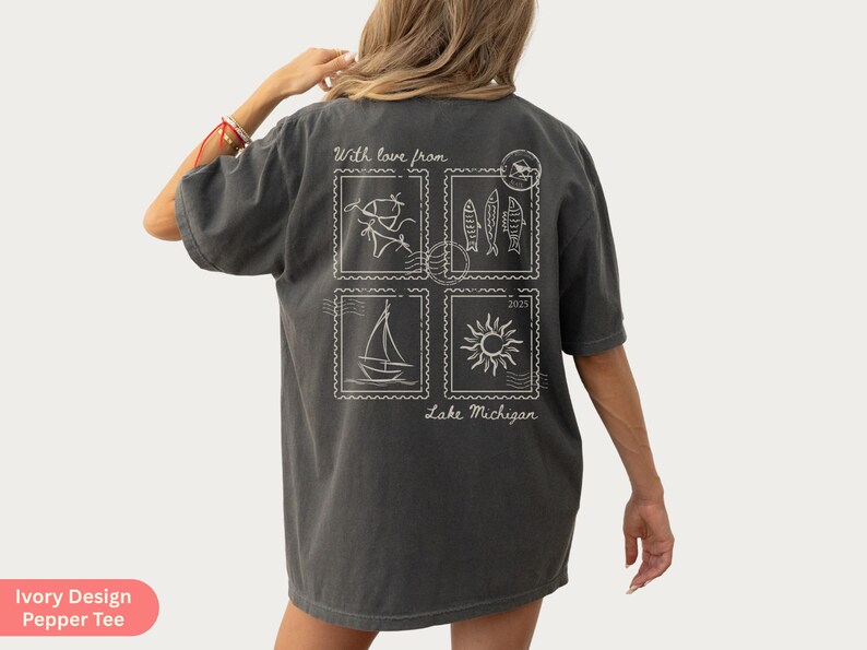 May include: Dark gray t-shirt with a white graphic design on the back. The design features four postage stamp-like images with a sailboat, fish, sun, and other elements. Text includes "With love from Lake Michigan 2023."