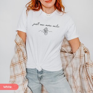 May include: White t-shirt with the text "just one more mile" in a script font, along with a line drawing of a running shoe. The shirt is paired with blue jeans and a plaid shirt.
