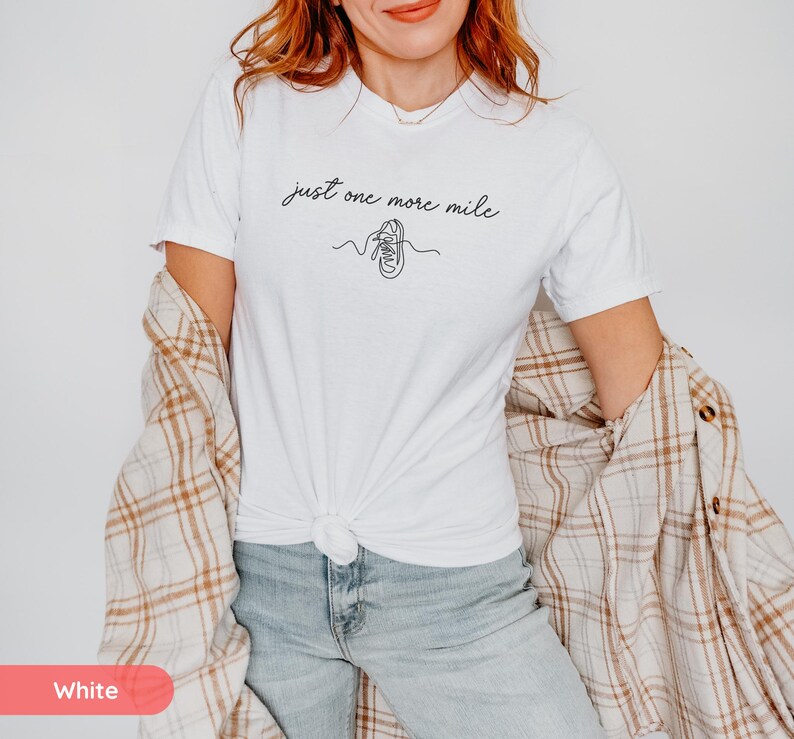 May include: White t-shirt with the text "just one more mile" in a script font, along with a line drawing of a running shoe. The shirt is paired with blue jeans and a plaid shirt.