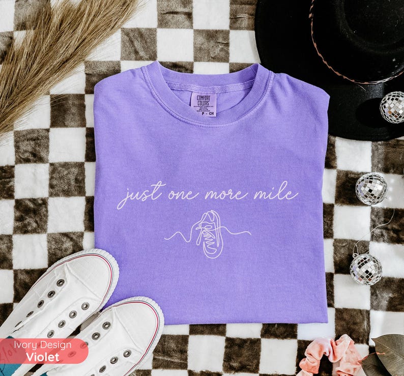 May include: A lavender t-shirt with the text "just one more mile" and a line drawing of a running shoe. White canvas sneakers are at the bottom left. The shirt is on a black and white checkered surface.