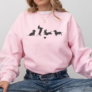 May include: A pink sweatshirt with a graphic of four dachshund silhouettes in various poses. The graphic includes a small heart.