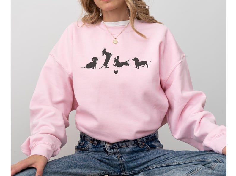 May include: A pink sweatshirt with a graphic of four dachshund silhouettes in various poses. The graphic includes a small heart.