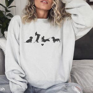 May include: A white sweatshirt with a graphic of four dachshunds in a line. The dachshunds are black silhouettes and are facing the right. The graphic includes a small heart between the second and third dachshund.