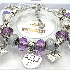 May include: A silver charm bracelet with purple and silver beads, a charm that says "Life is a Gift", and other charms including a teddy bear, a heart, and a scroll that says "Sweet".