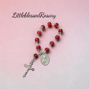 May include: A rosary featuring small, round, red crackle glass beads linked by silver-toned metal wire. It includes a silver-toned metal crucifix and an oval-shaped religious medal depicting a figure. The text LittleblessedRosary is visible.