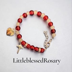 May include: A bracelet featuring alternating red and translucent off-white beads, a silver-toned crucifix charm, and a heart-shaped charm with an image. The text LittleblessedRosary is printed below the bracelet on a plain white background.