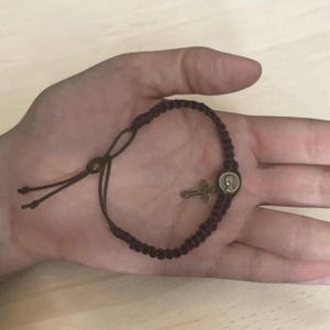 May include: A dark brown cord bracelet featuring a series of woven knots, a small circular metal charm depicting a profile of a figure, and a dangling ornate metal cross charm. The bracelet includes an adjustable sliding knot closure system.