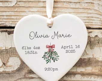 May include: White heart-shaped ceramic ornament with a mistletoe design, the text "Olivia Marie", "6 lbs 4 oz 18.5 in", "April 16 2025", and "9:20pm".