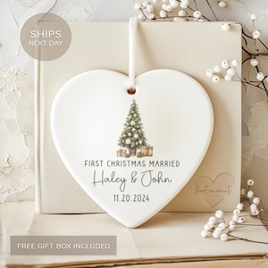 May include: A white heart-shaped Christmas ornament with a watercolor Christmas tree and gifts. Text reads "FIRST CHRISTMAS MARRIED" with names "Haley & John" and the date "11.20.2024". A white ribbon is attached for hanging.