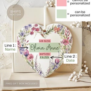 May include: A white ceramic heart-shaped ornament with a floral design. The ornament has the text "God Bless Olivia Anne Baptized 8.16.2024." on it. The ornament is on a book with a white lace overlay.
