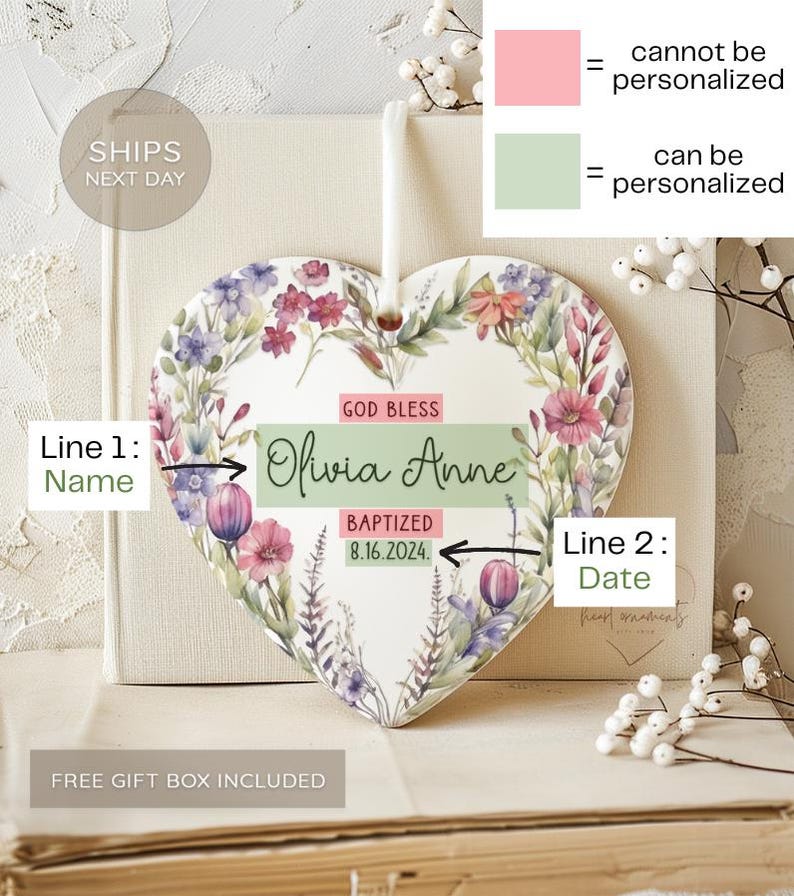 May include: A white ceramic heart-shaped ornament with a floral design. The ornament has the text "God Bless Olivia Anne Baptized 8.16.2024." on it. The ornament is on a book with a white lace overlay.