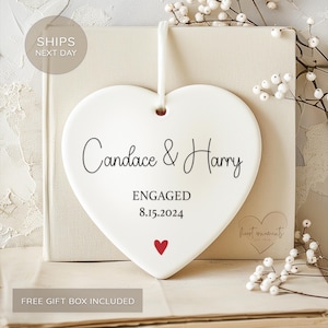 May include: White ceramic heart ornament with the text "Candace & Harry" and "ENGAGED 8.15.2024".