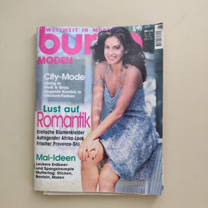 Puede incluir: Una portada de revista vintage con una mujer con un vestido floral azul. El título de la revista es "bur" en letras rosas grandes, con las palabras "MODEN" y "City-Mode" debajo. La portada también incluye texto en alemán.