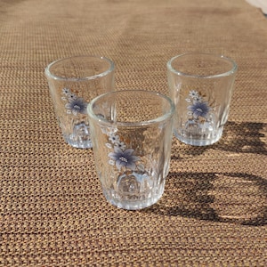 May include: Three clear glass shot glasses with a floral design. The flowers are blue and white with a yellow center.