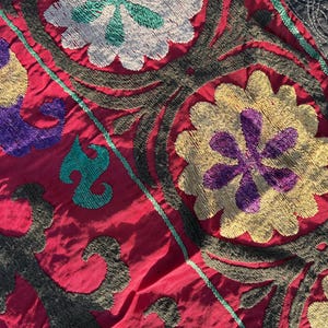 May include: Close-up of a vibrant textile with detailed floral embroidery. The design features a rich colour palette of red, purple, yellow, green, and grey. The detailed stitching creates a textured surface, highlighting the craftsmanship of the piece.
