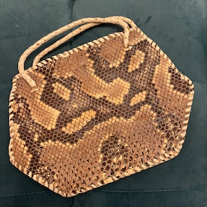 May include: A snakeskin handbag with a braided handle. The bag features a pattern of brown, tan, and black scales. The edges are stitched with a light-colored thread, and the handle is a cream color.