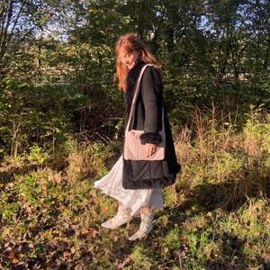 K&ouml;nnte beinhalten: Eine Frau tr&auml;gt einen schwarzen Mantel mit Kunstpelzbesatz und eine rosafarbene gesteppte Tasche &uuml;ber der Schulter. Sie steht in einem Waldgebiet mit heruntergefallenen Bl&auml;ttern auf dem Boden.