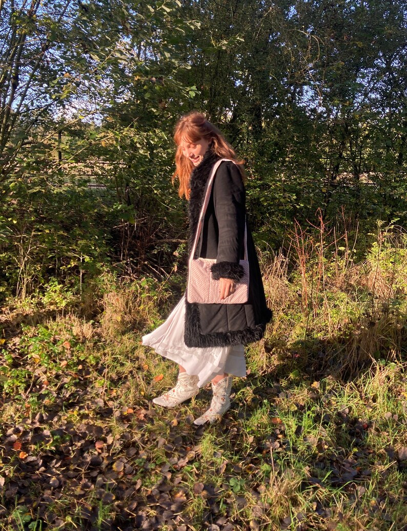 K&ouml;nnte beinhalten: Eine Frau tr&auml;gt einen schwarzen Mantel mit Kunstpelzbesatz und eine rosafarbene gesteppte Tasche &uuml;ber der Schulter. Sie steht in einem Waldgebiet mit heruntergefallenen Bl&auml;ttern auf dem Boden.