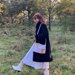 Może przedstawiać: Kobieta w czarnym płaszczu i białej sp&oacute;dnicy idzie przez trawiastą łąkę. Niesie patchworkową torbę w r&oacute;żowo-ż&oacute;łto-pomarańczowym wzorze. Ma na sobie białe kowbojskie buty.