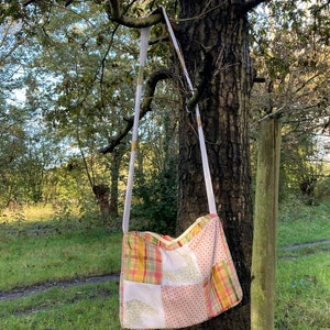 Może przedstawiać: Torba typu tote z patchworku z białym paskiem wiszącym na gałęzi drzewa. Torba jest wykonana z r&oacute;żnych tkanin w odcieniach r&oacute;żowego, ż&oacute;łtego i zielonego.