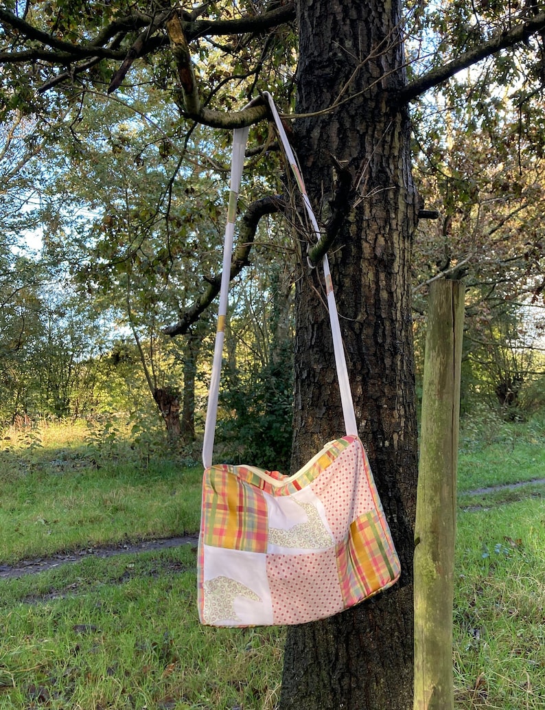 Może przedstawiać: Torba typu tote z patchworku z białym paskiem wiszącym na gałęzi drzewa. Torba jest wykonana z r&oacute;żnych tkanin w odcieniach r&oacute;żowego, ż&oacute;łtego i zielonego.