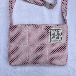 K&ouml;nnte beinhalten: Eine rosa-wei&szlig;e gepunktete Stoff-Umh&auml;ngetasche mit einem kleinen Patch, der eine Schwarzwei&szlig;illustration von zwei Figuren mit dem Text "franky+lucy" zeigt.