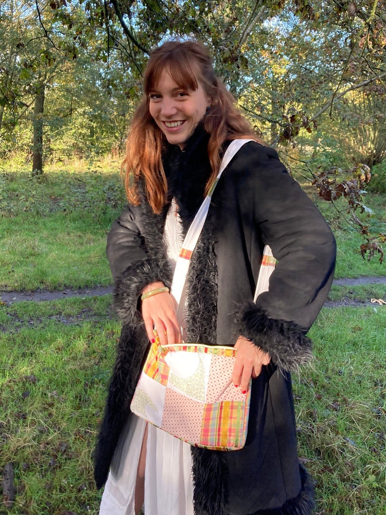 Może przedstawiać: Kobieta w czarnym płaszczu z imitacji futra z białą i kolorową patchworkową torbą przewieszoną przez ramię. Uśmiecha się i stoi na trawiastym terenie z drzewami w tle.