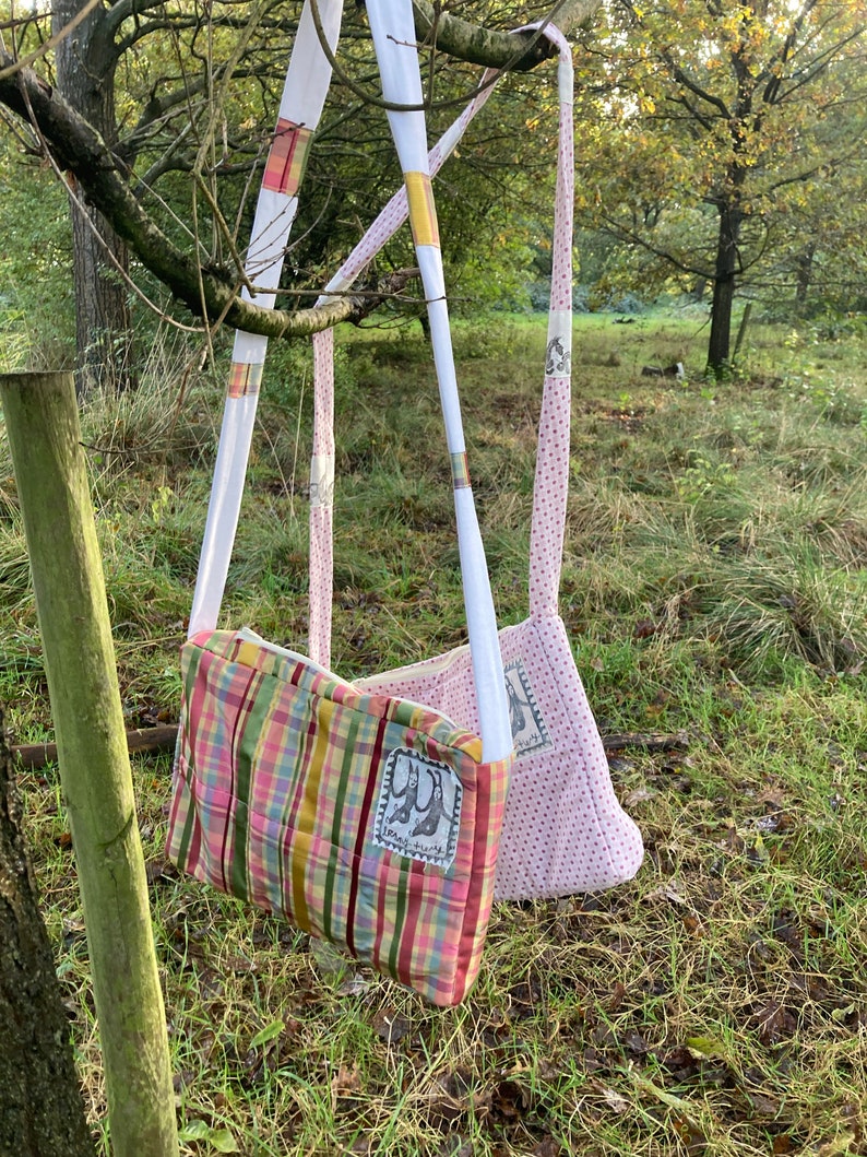 Może przedstawiać: Dwie torby materiałowe wiszące na gałęzi drzewa. Torba po lewej stronie jest patchworkiem z r&oacute;żowych, zielonych, ż&oacute;łtych i białych pask&oacute;w. Torba po prawej stronie jest r&oacute;żowa w białe kropki. Obie torby mają biały pasek i małą haftowaną naszywkę z wzorem dw&oacute;ch kr&oacute;lik&oacute;w.