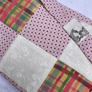 K&ouml;nnte beinhalten: Ein Patchwork-Deckenst&uuml;ck mit einem rosa-wei&szlig;en Polka-Dot-Stoff, einem wei&szlig;en Stoff mit Blumenmuster, einem karierten Stoff in den Farben Rosa, Gr&uuml;n und Gelb und einem wei&szlig;en Stoff mit einer schwarz-wei&szlig;en Illustration von zwei Drachen.