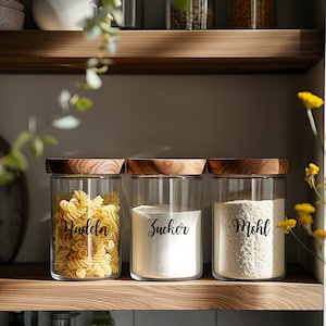 Könnte beinhalten: Drei Glasbehälter mit Holzdeckeln und Etiketten. Die Behälter sind mit Nudeln, Zucker und Mehl gefüllt. Die Etiketten lauten "Nudeln", "Zucker" und "Mehl".