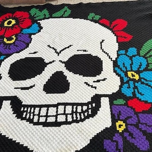 Puede incluir: Una manta de ganchillo negra que presenta una calavera blanca grande centrada en el diseño. La calavera está rodeada de coloridos acentos florales en tonos rojo, azul, morado y verde, creados con un patrón de punto texturizado y pixelado.