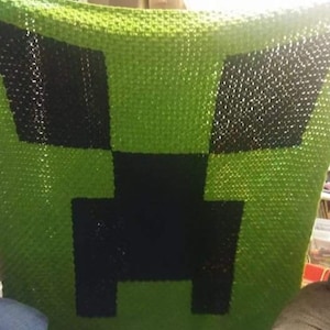 May include: A handmade, green and black crocheted blanket with a pixelated design. The blanket's pattern resembles a character from a popular video game. The blanket is made of soft yarn and is suitable for use as a throw or decorative piece.