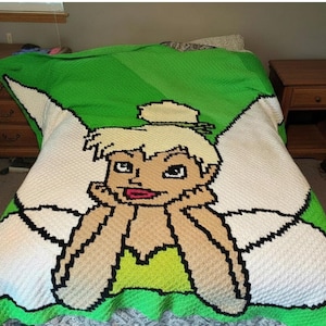 Puede incluir: Una manta tejida a ganchillo a mano que presenta un gran gráfico central de un personaje de hada rubia con un moño alto, apoyando la barbilla en las manos. El diseño utiliza hilo blanco, beige y negro sobre un fondo verde vibrante con alas.
