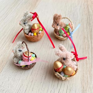 Puede incluir: Cuatro cestas de Pascua en miniatura, cada una con un conejito de peluche, huevos coloridos y caramelos. Las cestas son tejidas y cada una tiene una cinta para colgar. Los conejitos son en tonos grises y marrones, y los huevos son dorados, rosas y verdes.
