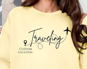 Könnte beinhalten: Ein gelber Pullover mit dem Text "Traveling" in schwarzer Kursivschrift. Ein kleines Flugzeugsymbol und ein Pin-Symbol mit dem Text "Custom Location" sind ebenfalls auf dem Pullover aufgedruckt.