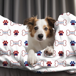 Puede incluir: Una cama para perro blanca con un diseño de huella de pata y hueso rojo y azul. La cama tiene el nombre "Hagrid" impreso. Un perro marrón y blanco asoma por debajo de la cama.