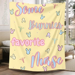 Puede incluir: Una manta amarilla con el texto "Some Bunnies Favorite Nurse" en rosa, azul y verde. La manta está decorada con conejos y símbolos médicos.