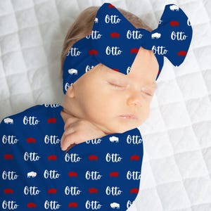 May include: A baby wearing a navy blue swaddle and matching bow headband. The items are patterned with white bison and the name "Otto" in white, and red bison. The baby is lying on a white quilted surface.