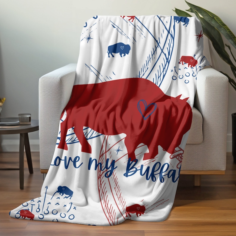 Peut inclure: Une couverture en polaire blanche avec une silhouette de bison rouge et le texte "Love my Buffalo". La couverture a un motif bleu et blanc avec des &eacute;toiles et des silhouettes de bisons.