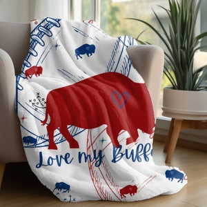 Peut inclure: Jet&eacute; en polaire blanc avec une silhouette de buffle rouge et le texte "Love my Buff" en bleu. Le jet&eacute; pr&eacute;sente un motif r&eacute;p&eacute;t&eacute; de silhouettes de buffles bleus, de c&oelig;urs rouges et bleus et de coutures de baseball rouges et bleues.
