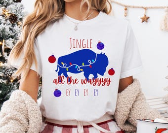 May include: White t-shirt with a festive design. The shirt features a blue buffalo silhouette adorned with Christmas lights and the words "Jingle all the wayyyy." Red and blue ornaments complete the holiday theme. A cozy, seasonal apparel item.