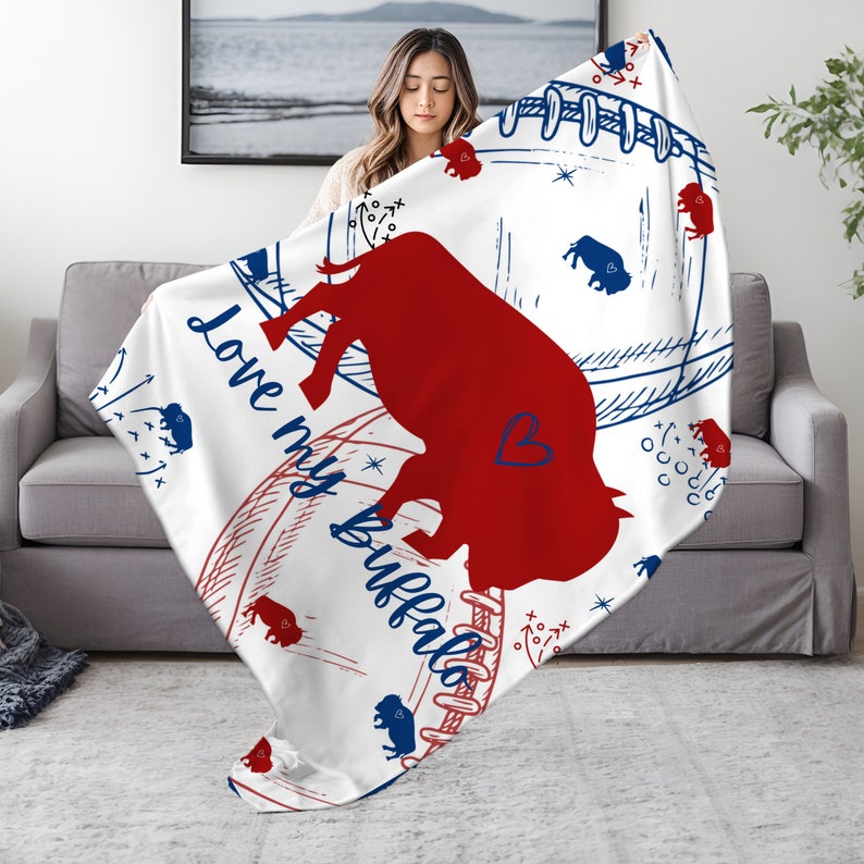 Peut inclure: Une couverture en polaire blanche avec une silhouette de bison rouge et le texte "Love my Buffalo" en bleu. La couverture pr&eacute;sente un motif de football bleu et blanc avec des &eacute;toiles et d'autres &eacute;l&eacute;ments graphiques.