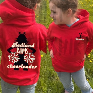 Könnte beinhalten: Roter Kapuzenpullover mit Vordertasche und Grafik auf dem Rücken. Die Grafik auf der Rückseite zeigt eine Cheerleader-Silhouette, Pompons und die Worte "Redland RAMS cheerleader". Auf der Vorderseite befindet sich eine Cheerleader-Silhouette und der Name "Veronica".