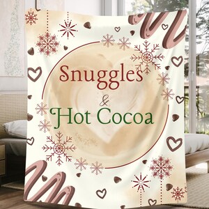 Może przedstawiać: Kremowy koc z motywem kawowym. Koc przedstawia duży wzór filiżanki kawy ze słowami "Snuggles & Hot Cocoa" w kolorze czerwonym i zielonym. Ozdobne płatki śniegu i serca są rozrzucone wokół wzoru.