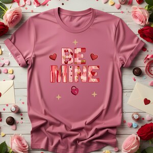 May include: A pink t-shirt with the words "BE MINE" in a decorative font. The letters are filled with patterns of hearts and floral designs. The shirt also features heart and star accents, and a heart-shaped lock.