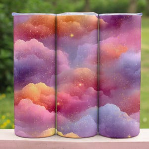 May include: Three stainless steel tumblers with a vibrant, colorful design. The tumblers feature a dreamy, cloud-like pattern in shades of pink, purple, orange, and yellow, with sparkling accents. The tumblers are cylindrical and have a lid.