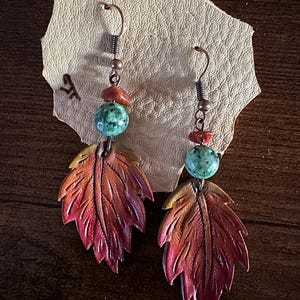 May include: A pair of leather earrings shaped like autumn leaves with a red, orange, and yellow gradient. Each earring features a turquoise and red bead.