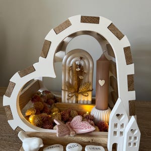 Könnte beinhalten: Ein weißer Holzrahmen in Herzform mit einer Kerze, getrockneten Blumen und einem rosa Schmetterling. Der Rahmen ist mit Holzapplikationen verziert und trägt die Worte "Freude", "Glück" und "Liebe" auf der Basis.